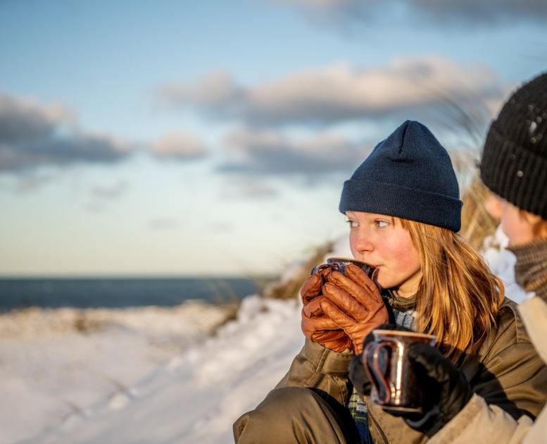 Jente holder varmen med en kopp varm drikke ved Klitmøller strand om vinteren