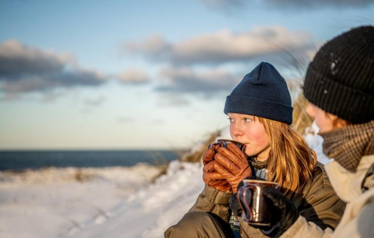 Jente holder varmen med en kopp varm drikke ved Klitmøller strand om vinteren